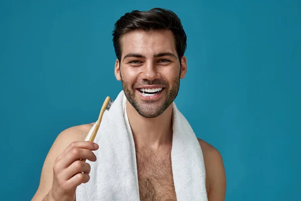 Portrait of happy bearded man with towel around neck smiling at camera with toothpaste in his mouth, holding wooden toothbrush isolated over blue background — Stock Photo, Image