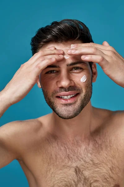 Cheerful good looking guy smiling at camera while applying white cream on his face, posing isolated over blue background — Stock Photo, Image