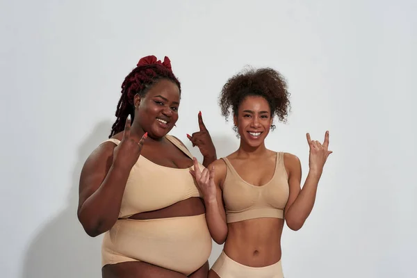 Dos mujeres afroamericanas sonrientes en ropa interior con diferentes tamaños de cuerpo haciendo gestos de rock and roll, posando juntas aisladas sobre un fondo gris —  Fotos de Stock