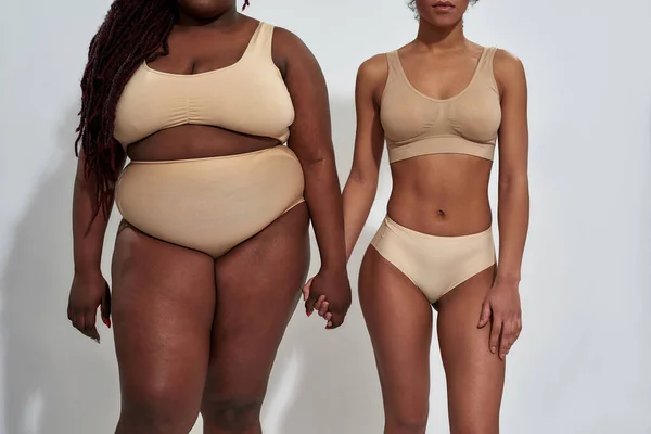 Gehakte foto van twee Afrikaanse Amerikaanse vrouwen in ondergoed met verschillende lichaamsgrootte die elkaars handen vasthouden, die samen geïsoleerd staan over een grijze achtergrond — Stockfoto