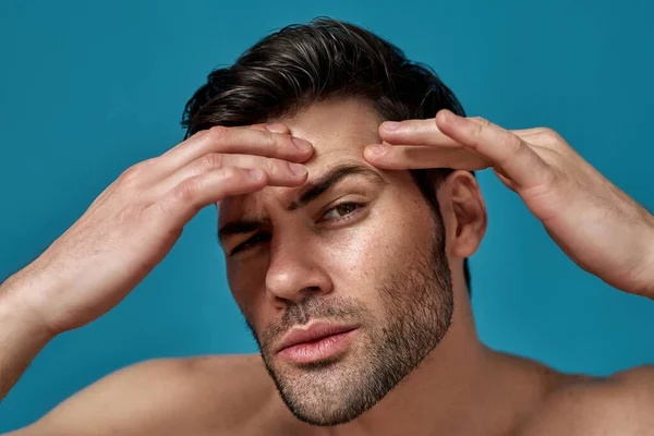 Close-up portret van shirtloze knappe man controleren van zijn huid, op zoek naar vlekken terwijl poseren geïsoleerd over blauwe achtergrond — Stockfoto