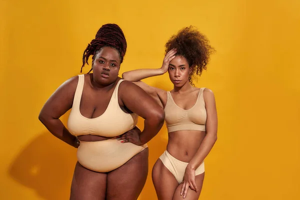 Studio shot van twee zelfverzekerde plus size en slanke Afrikaanse Amerikaanse vrouwelijke modellen in beige ondergoed kijken naar de camera terwijl samen poseren geïsoleerd over gele achtergrond — Stockfoto