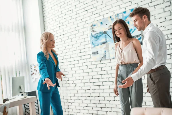 Pareja joven visitando agencia inmobiliaria para comprar, alquilar piso o casa. Mujer encantadora dando la bienvenida a la nueva familia en la oficina moderna —  Fotos de Stock