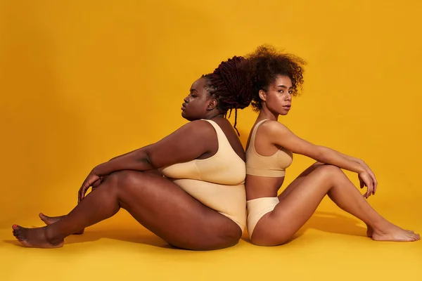 Volledige lengte schot van twee diverse Afrikaanse Amerikaanse vrouwelijke modellen in ondergoed zitten samen geïsoleerd over gele achtergrond — Stockfoto