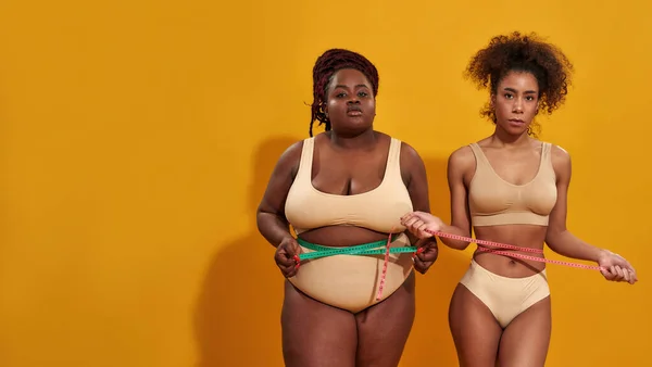 Twee jonge Afro-Amerikaanse vrouwen met verschillende lichaamsgrootte kijken naar camera, meten hun taille met metrische meetlint, staan samen geïsoleerd op oranje achtergrond — Stockfoto