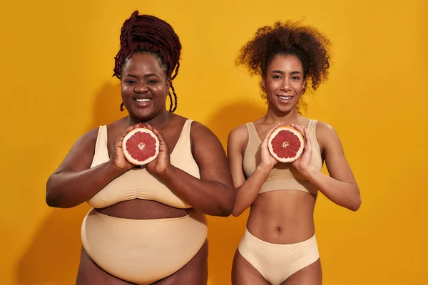Estudio de dos alegres mujeres afroamericanas en ropa interior sonriendo a la cámara, sosteniendo mitades de jugoso pomelo frente a ellas, posando aisladas sobre fondo naranja — Foto de Stock