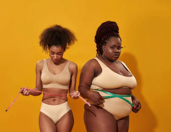 Twee Afro-Amerikaanse vrouwen met verschillende lichaamsvorm meten hun taille met metrische meetlint, staan samen geïsoleerd op oranje achtergrond — Stockfoto