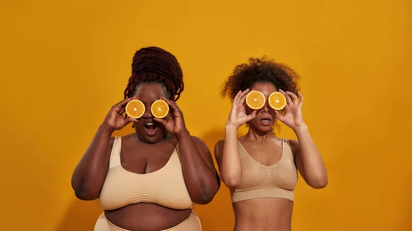 두 명의 아프리카 계 미국인 여성 이 속옷 차림으로, 잘 익은 즙 이 많은 오렌지 반쪽으로 눈을 가리고, 오렌지 색 배경 위로 고립된 채 포즈를 취하고 있다 — 스톡 사진