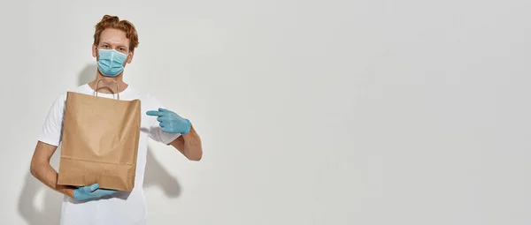 Obrázek mladého zrzavého kurýra držícího papírový sáček a ukazujícího na něj prstem — Stock fotografie