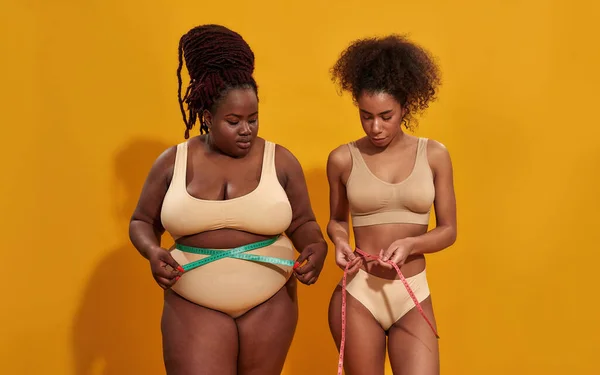 Dos jóvenes afroamericanas con diferente tamaño corporal midiendo su cintura usando cinta métrica después de una dieta, posando juntas aisladas sobre fondo naranja — Foto de Stock