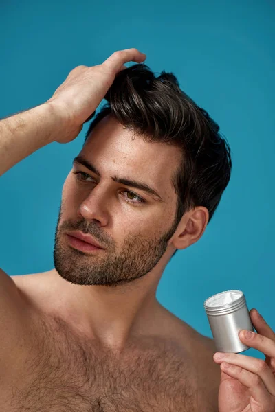 Closeup portrait of sexy brunette guy with bristle holding silver jar with gel or cream and applying it on his dark brown clean hair isolated over blue background — Stock Photo, Image