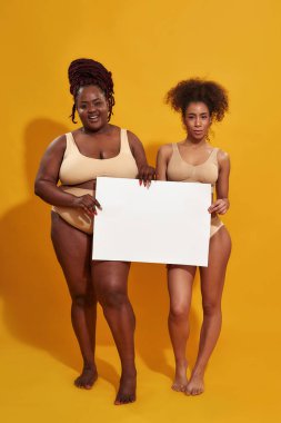 İç çamaşırlı, farklı vücut şekline sahip iki Afro-Amerikan kadının kameraya boş beyaz pankartla baktığı, sarı arka planda tek başına durduğu uzun bir çekim.