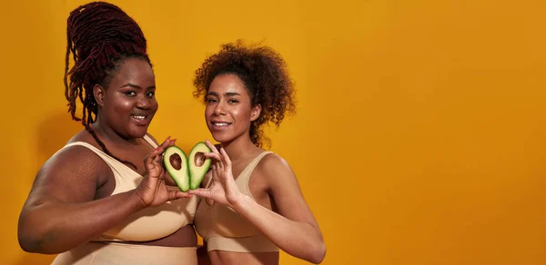 Dos mujeres afroamericanas en ropa interior sonriendo ante la cámara, sosteniendo mitades de aguacate de frutas saludables como un signo del corazón, posando aisladas sobre un fondo amarillo — Foto de Stock