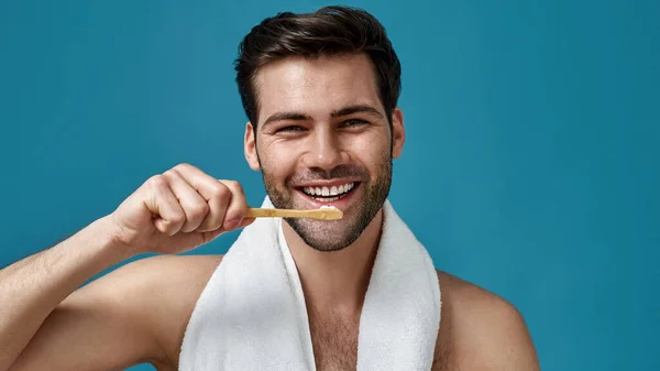 Beauty portret van goed uitziende brunette kerel met handdoek om zijn nek glimlachen op camera, vasthouden tandenborstel en poetsen zijn tanden terwijl poseren geïsoleerd over blauwe achtergrond — Stockfoto