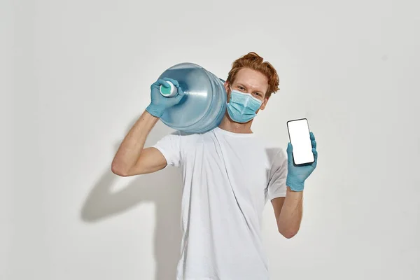 Imagem de um jovem entregador magro carregando uma enorme garrafa de água e anunciando um aplicativo — Fotografia de Stock