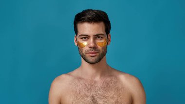 Mavi arka planda izole edilmiş kameraya bakan altın rengi göz bantları olan beyaz bir adamın stüdyo portresi.