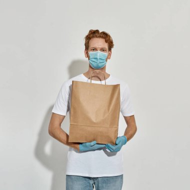 Genç kızıl saçlı adam maske takıyor ve elinde bir kese kağıdı tutuyor.