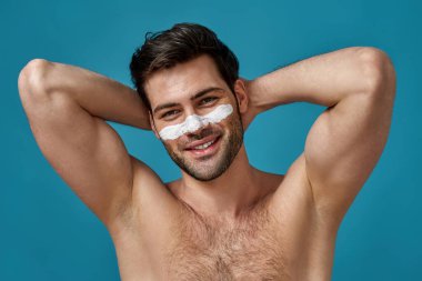 Beyaz maskeli neşeli esmer adam kameraya gülümseyen gözenekleri temizlemek için burnuna sürtünmüş, mavi arka planda izole edilmiş poz vermiş.