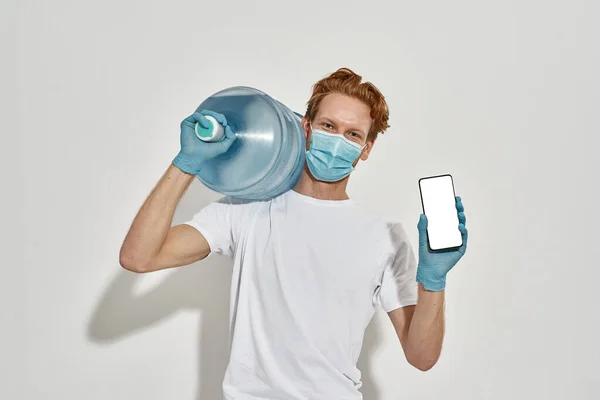 A kép egy fiatal vörös hajú futár kezében egy hatalmas üveg vizet, és gazdaság egy okostelefon — Stock Fotó