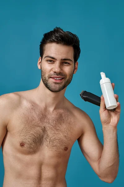 Beauty portrait of good looking brunette guy smiling at camera, holding electric shaver and shaving foam for better shave, to protect skin from irritation isolated over blue background — Stock Photo, Image