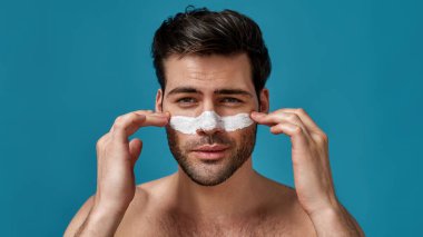 Kahverengi saçlı ve beyaz maskeli çekici bir adam kameraya bakarak gözenekleri temizlemek için burnuna sürtünmüş ve mavi arka planda izole bir şekilde poz vermiş.