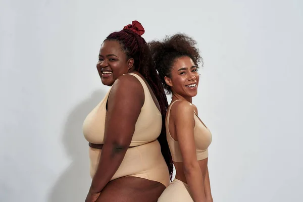 Dos mujeres afroamericanas alegres en ropa interior con forma de cuerpo diferente de pie espalda con espalda, sonriendo a la cámara aislada sobre fondo gris — Foto de Stock