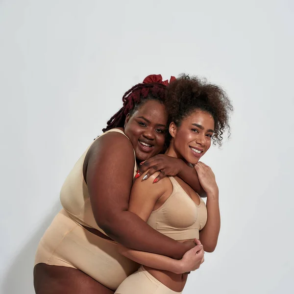 Dos mejores amigas, mujeres afroamericanas con curvas y delgadas en ropa interior abrazando y sonriendo a la cámara, posando juntas aisladas sobre un fondo gris — Foto de Stock