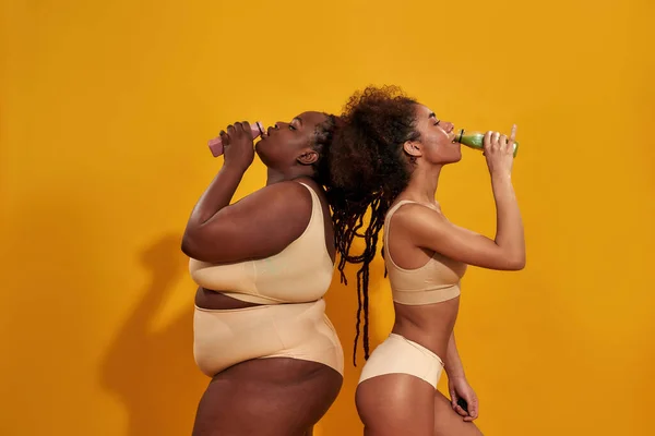 Side view of two young african american women with different body size standing back to back and drinking smoothie, posing together isolated over orange background — Stock Photo, Image