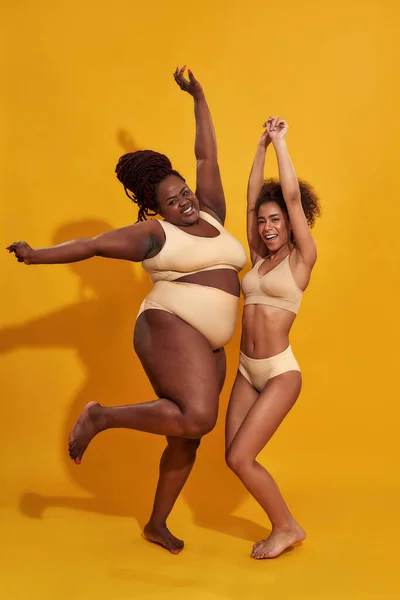 Foto cuerpo entero de dos mujeres afroamericanas en ropa interior beige diferentes tamaños de cuerpo se ven felices, juntas mientras posan aisladas sobre fondo amarillo: fotografía de stock ©