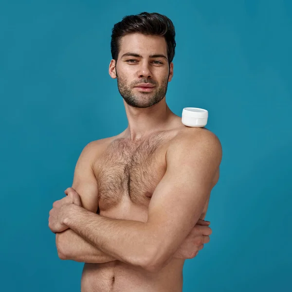 Studio shot of handsome naked brunette guy posing with white jar of effective cream on his shoulder, looking at camera, standing with arms crossed isolated over blue background — Stock Photo, Image