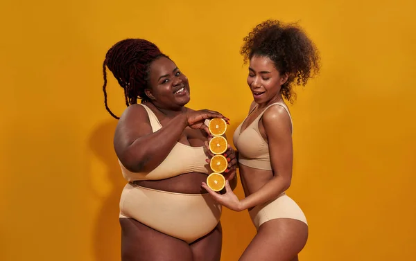 Studio girato di due donne afro-americane in biancheria intima sorridente mentre tengono in mano metà di arancio maturo succoso, in piedi isolato su sfondo arancione — Foto Stock