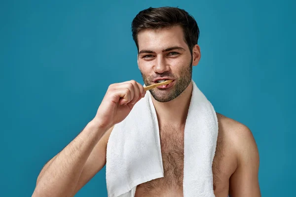Portret nagiego brunetka mężczyzna z ręcznikiem na szyi patrząc w aparat fotograficzny podczas mycia zębów odizolowany na niebieskim tle — Zdjęcie stockowe