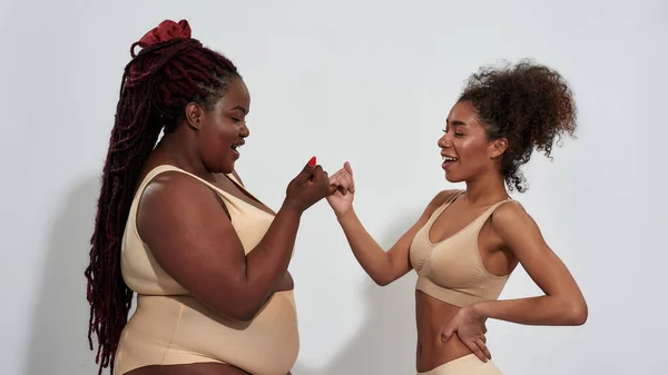Dos alegres mujeres afroamericanas en ropa interior con diferentes tamaños de cuerpo riendo, haciendo una promesa de meñique, de pie aislada sobre fondo gris — Foto de Stock