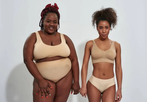 Dos mujeres afroamericanas en ropa interior beige con diferentes formas de cuerpo mirando a la cámara, de pie juntas aisladas sobre fondo gris —  Fotos de Stock