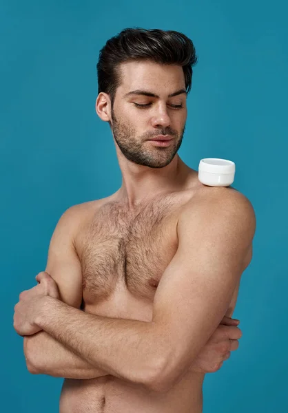 Studio shot van naakte brunette man op zoek naar witte pot effectieve crème op zijn schouder, poseren met armen gekruist geïsoleerd over blauwe achtergrond — Stockfoto