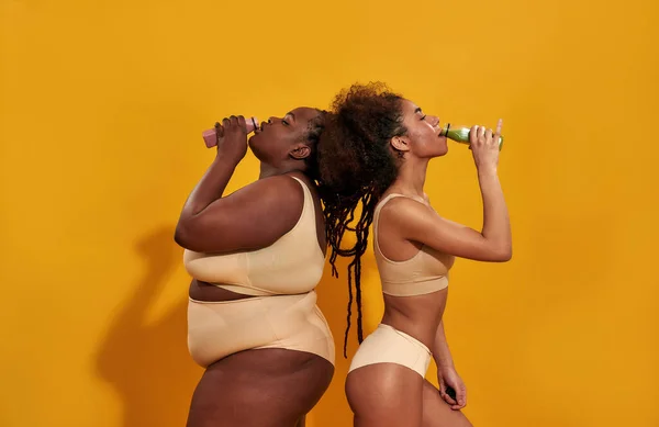 Foto cuerpo entero de dos mujeres afroamericanas en ropa interior beige diferentes tamaños de cuerpo se ven felices, juntas mientras posan aisladas sobre fondo amarillo: fotografía de stock ©