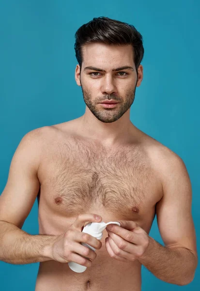 Handsome brunette naked man doing morning routine, applying foam beauty product on a cotton pad for cleansing his face, posing isolated over blue background — Stock Photo, Image