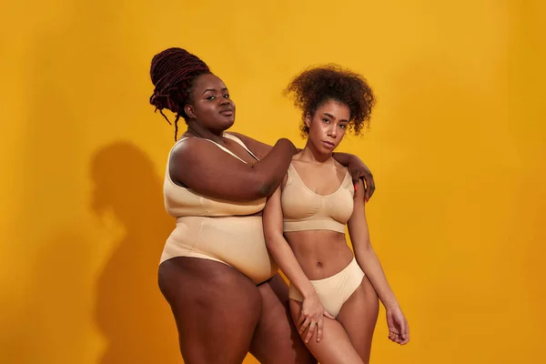 Dos mujeres afroamericanas serias en ropa interior beige con diferentes tamaños de cuerpo de pie juntas, posando aisladas sobre fondo amarillo — Foto de Stock