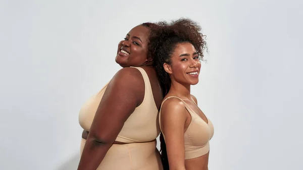 Due donne africane americane in biancheria intima con diversa forma del corpo in piedi schiena a schiena, sorridendo alla fotocamera isolata su sfondo grigio — Foto Stock