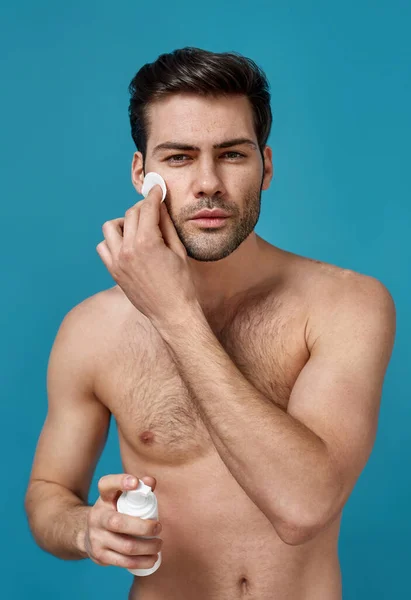 Sexy brunette naakte man doet ochtend routine, het reinigen van zijn gezicht met katoen pads voor het scheren en houden schuim reinigingsmiddel, poseren geïsoleerd over blauwe achtergrond — Stockfoto