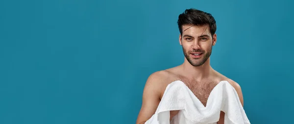 在演播室里描绘的是一个快乐的黑发男人，刮胡子和洗澡后，用白色毛巾擦拭自己，对着相机微笑，在蓝色的背景上摆出孤立的姿势 — 图库照片