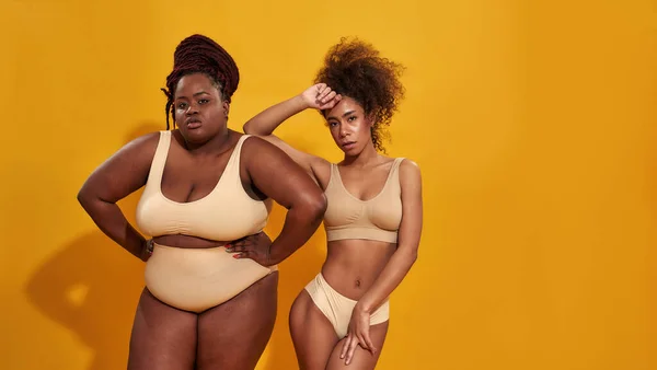 Estudio de dos mujeres afroamericanas seguras de más tamaño y delgadas en ropa interior beige mirando a la cámara mientras posan juntas aisladas sobre fondo amarillo —  Fotos de Stock