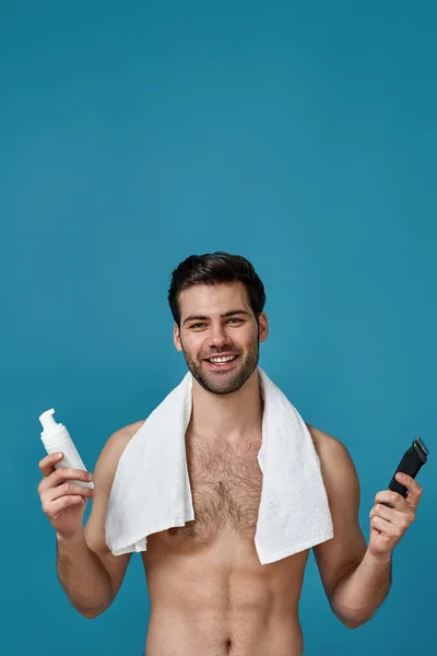 Nice looking naked guy with white towel around his neck smiling at camera, posing with shaving foam and electric shaver isolated over blue background — Stock Photo, Image
