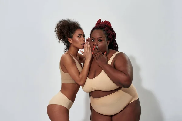 Mujer afroamericana delgada en ropa interior compartiendo un secreto con su amiga de talla grande, de pie juntos aislados sobre un fondo gris —  Fotos de Stock