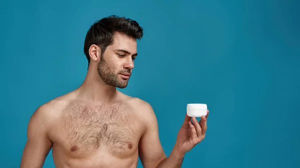 Portrait of naked model guy holding white jar with effective cream for face, posing isolated over blue background — Stok Foto