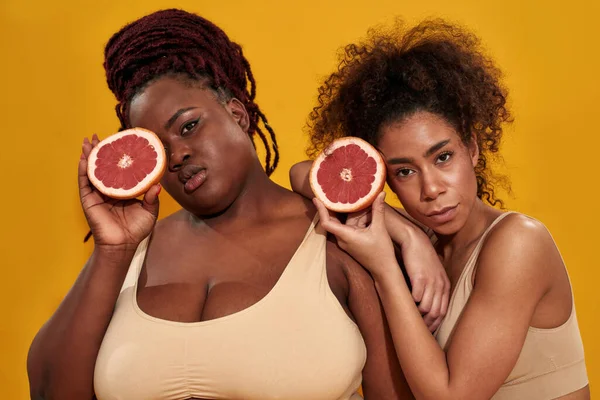 Две молодые африканские американки в нижнем белье смотрят в камеру, держат половинки сочного грейпфрута, позируют вместе изолированно на оранжевом фоне — стоковое фото