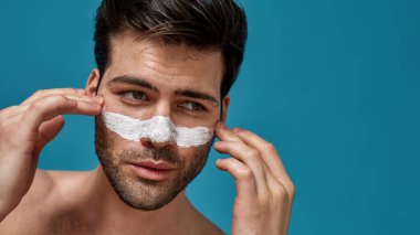 Yüzüne dokunan gözenekleri temizlemek için beyaz maskeli, temiz ciltli yakışıklı sakallı adamın stüdyo portresi.