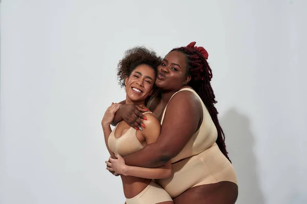 Two playful, curvy and slim african american women in underwear hugging while posing together for camera isolated over gray background — Stock Photo, Image