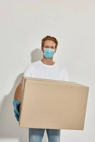 Imagen de un joven repartidor pelirrojo sosteniendo una enorme caja de cartón sobre la pared blanca — Foto de Stock