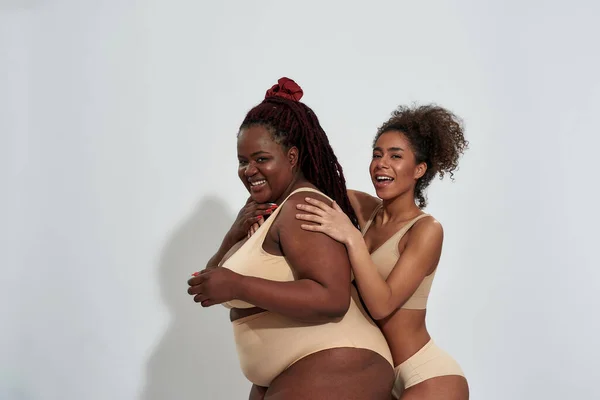 Fröhliche schlanke und übergewichtige Afroamerikanerinnen in beiger Unterwäsche lächeln, während sie isoliert vor grauem Hintergrund zusammen posieren — Stockfoto